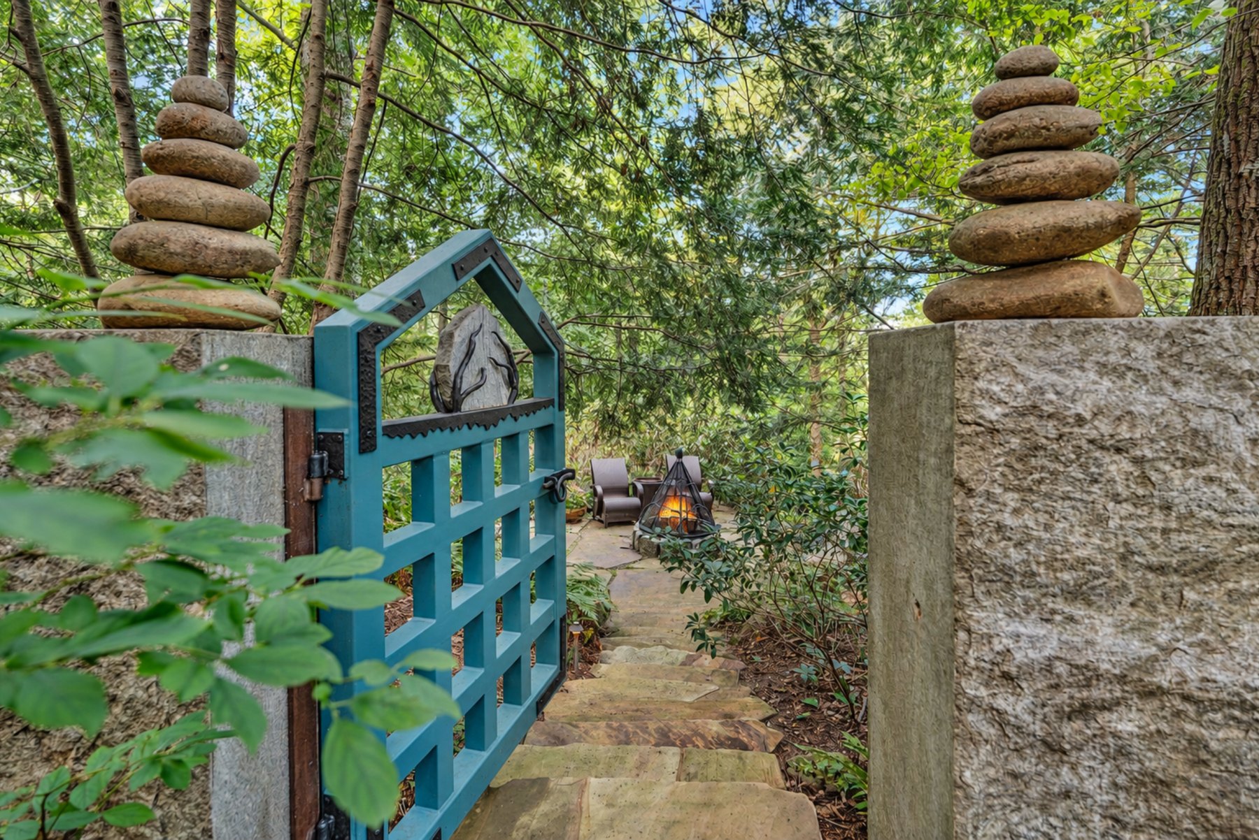 The blue gate standing open between two stone cairns, a flagstone path leading to the fire pit clearing